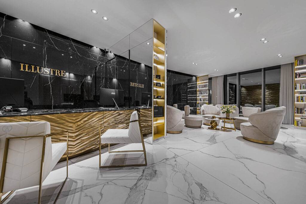 Nowoczesna, luksusowa recepcja z czarnymi i złotymi akcentami, marmurowymi podłogami, białymi krzesłami, okrągłymi beżowymi fotelami, regałami i dużymi oknami. Idealna dla osób poszukujących medycyny estetycznej Wrocław; ILLUSTRE i EXCELLIA na czarnej ścianie.